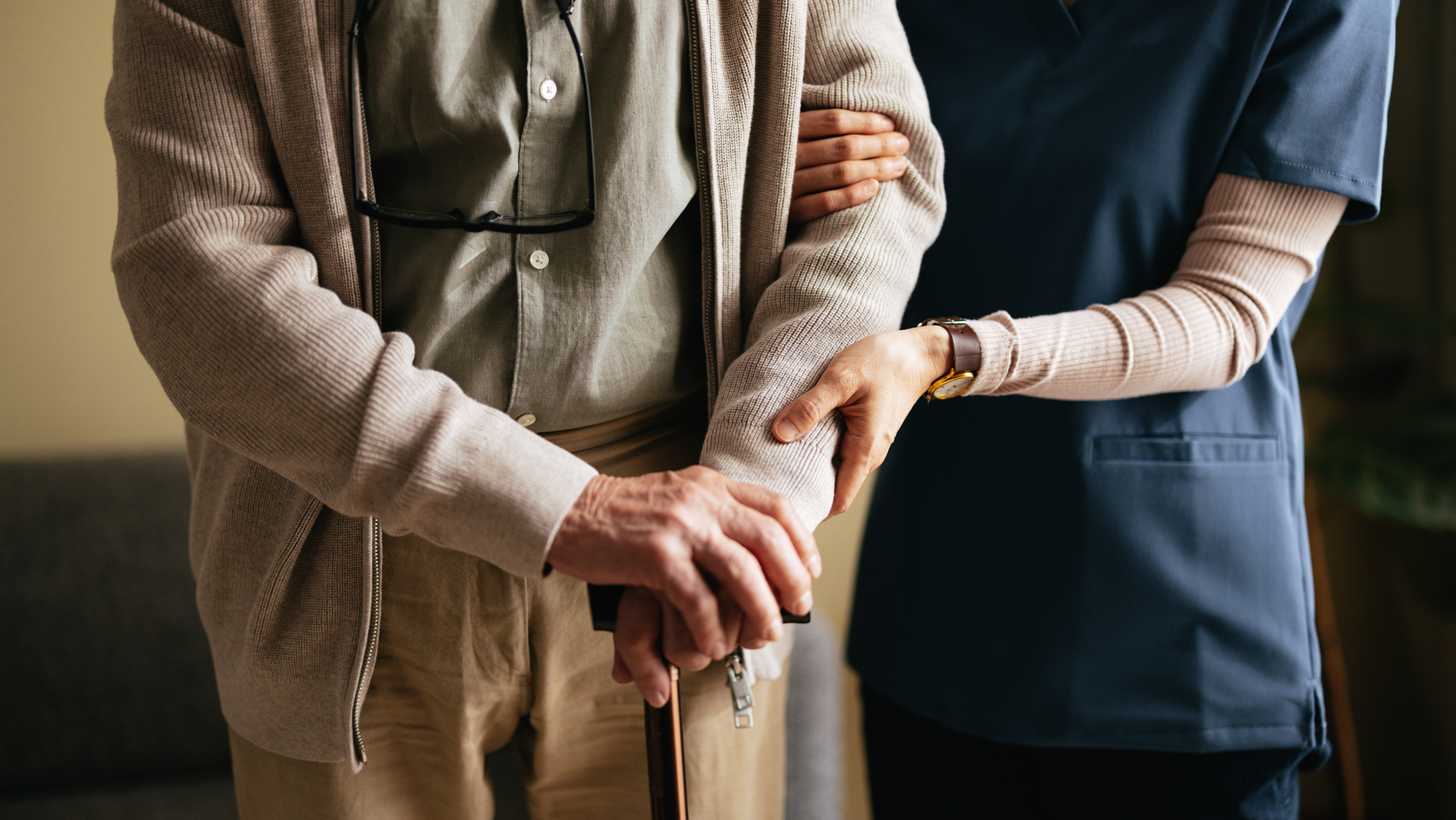 An older man holding a walking cane with a care provider holding his arm and standing beside him.