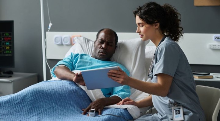 Image shows a man in a hospital bed looking at health information on a tablet with a medical professional.