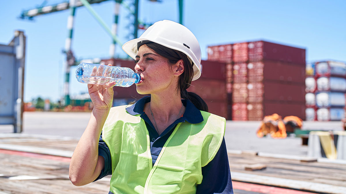 Female construction wearing a white hard hat and yellow safety vest, drinking water while working outside