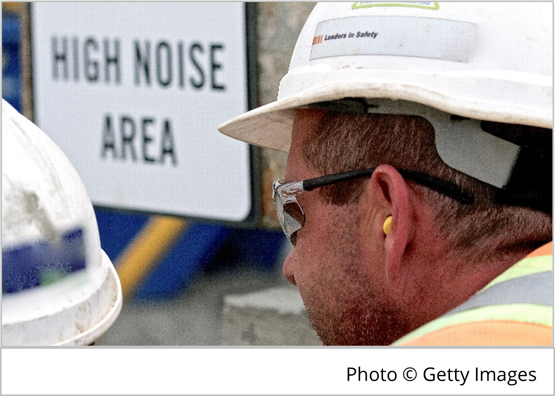 photo of a worker wearing ear plugs and a hard hat near a sign that says high noise area.