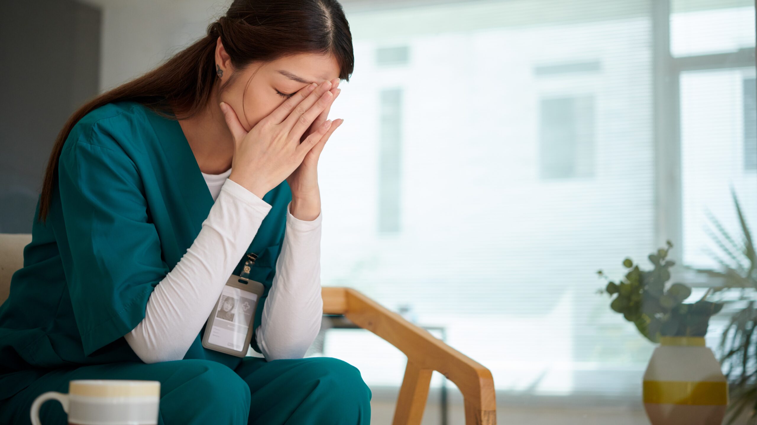 Female healthcare worker sitting down in a chair with her head in her hands, looking tired, with a mug next to her