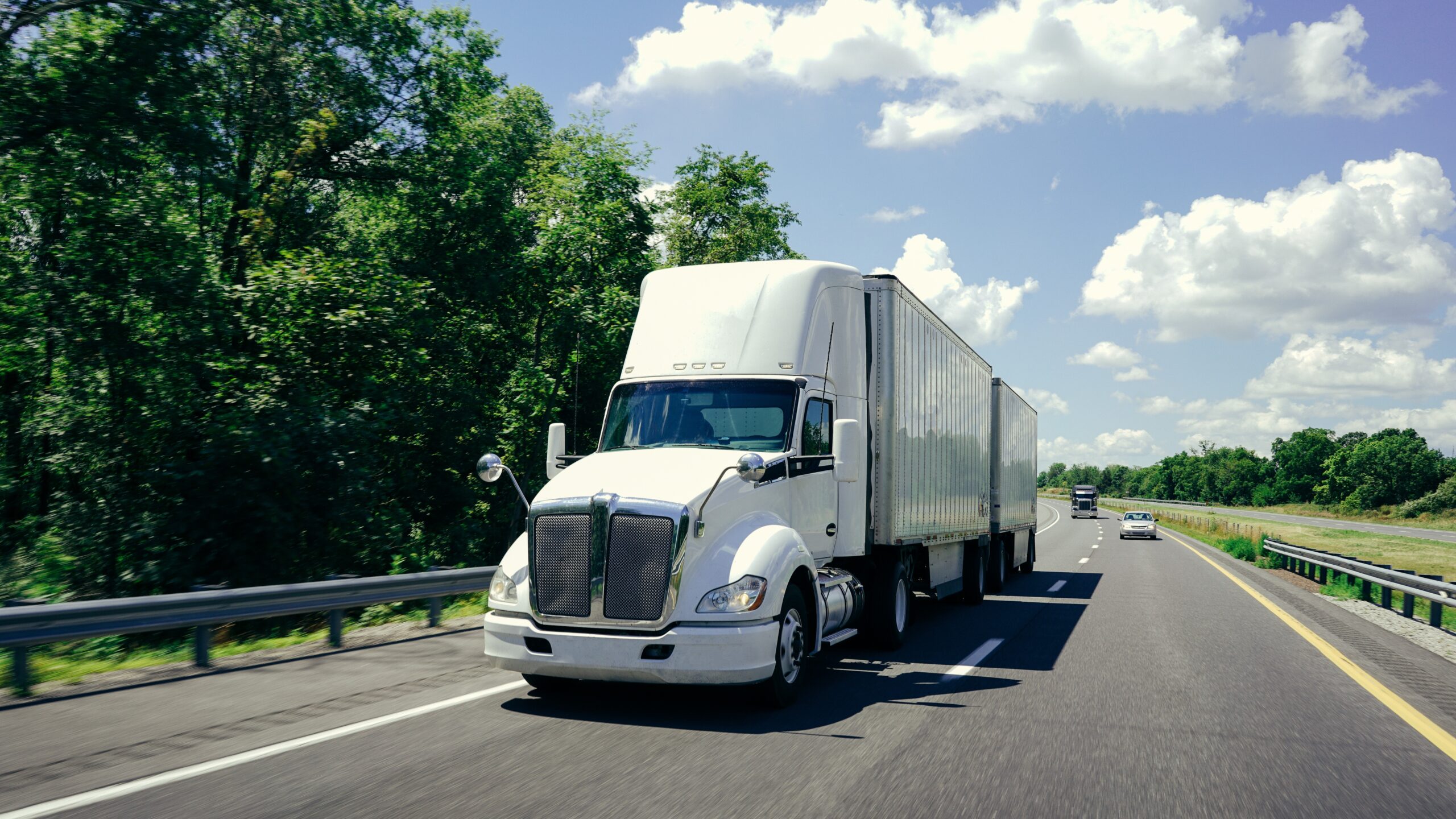 White eighteen wheeler truck on a tree-lined road, with blue sky and clouds above