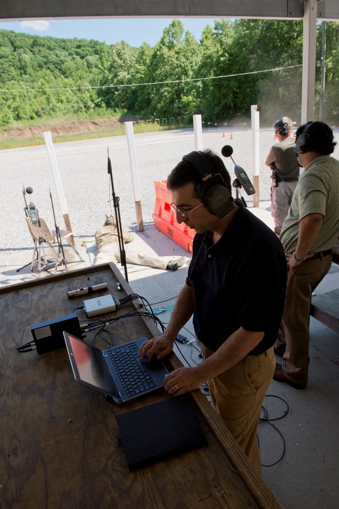 Figure 5: NIOSH researcher running the NIOSH impulse noise measurement system during live-fire training exercise (shown in the background are microphones positioned on tripods at ear level and prone positions).