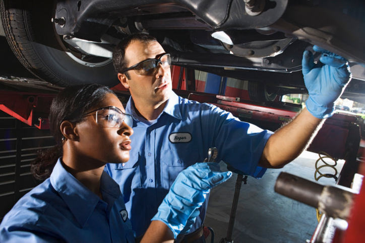 Two mechanics, a man and a woman wearing blue shirts, examine the underside of a car.