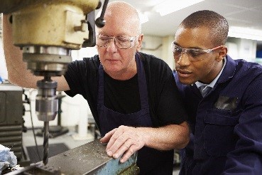 older white man with white hair showing a younger black man how to operate a piece of heavy machinery for craftsman work