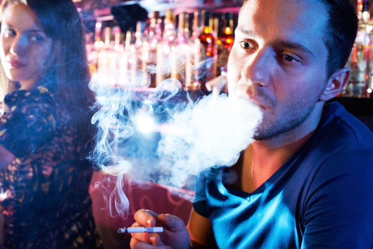White man smoking in a bar, with a white woman looking over her shoulder at him