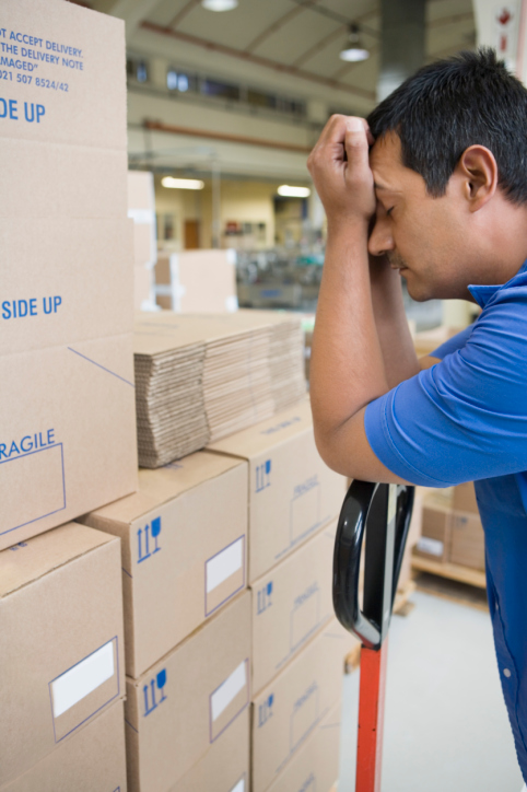 A tired male warehouse worker rests his head in his hands.