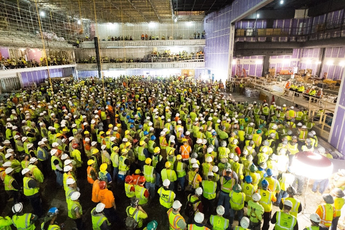 Photo of a room full of construction workers in hard hats and yellow vests.