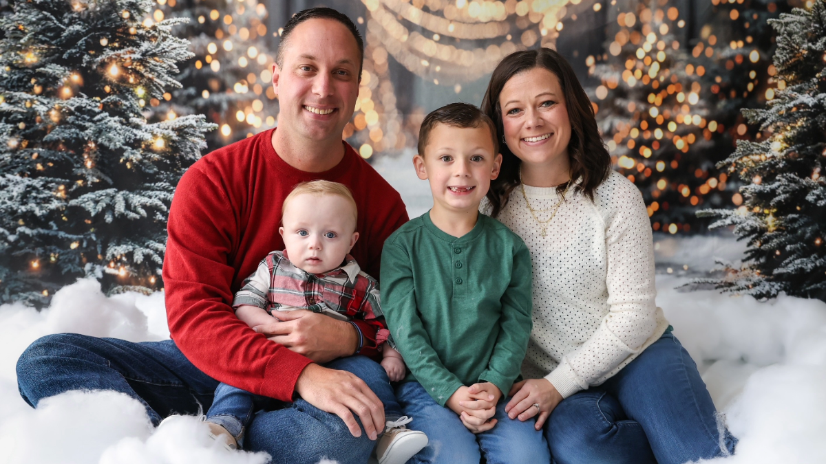 Two parents posing in front of Christmas tree backdrop with two small children