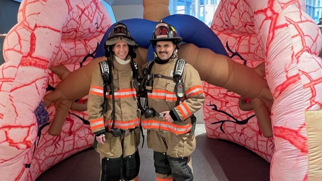 Two people dressed in firefighter turnout gear in front of inflatable lung display