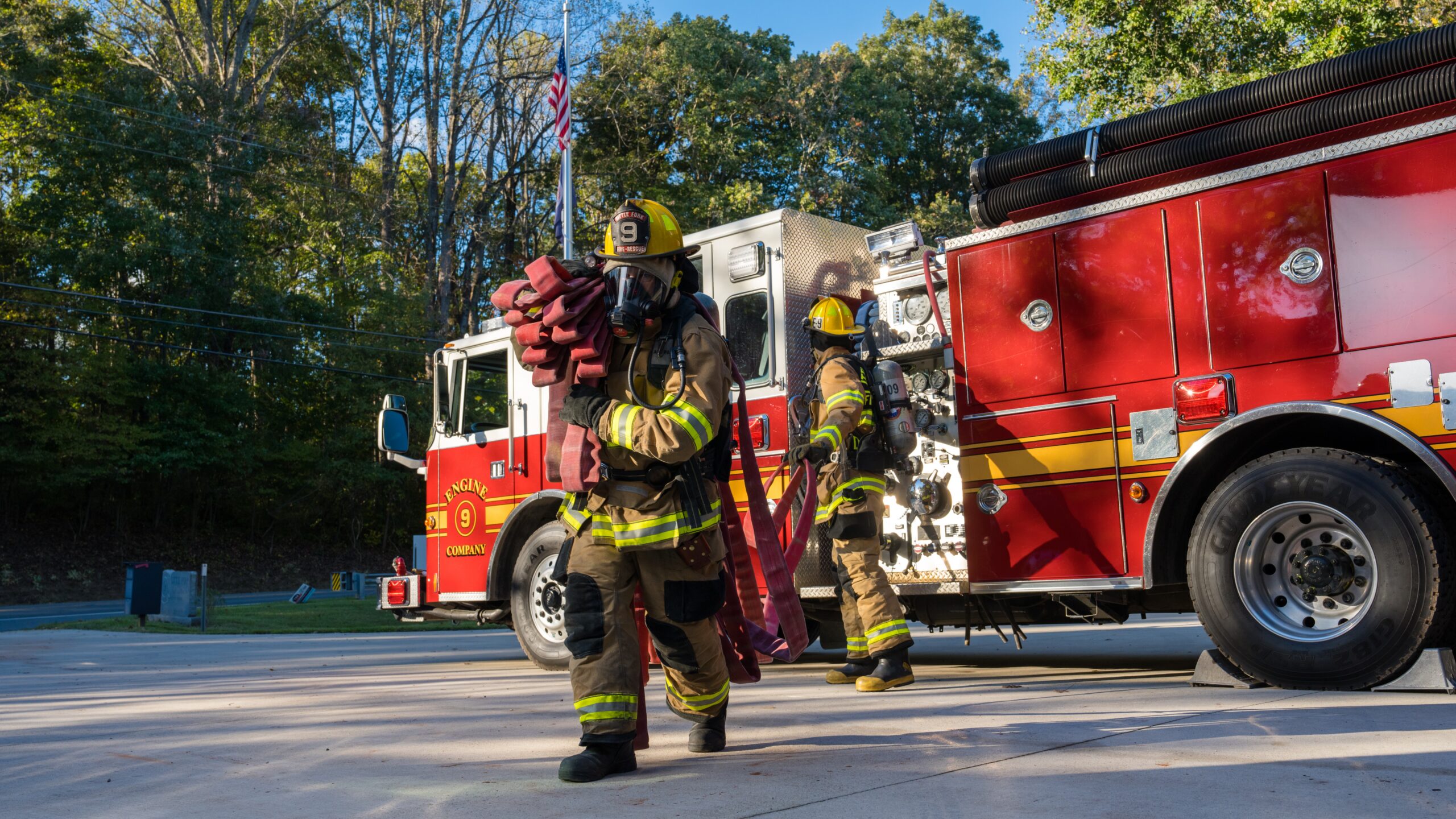 A firefighter carries a hoseline. As he gets further away from the truck another firefighter feeds more hoseline.