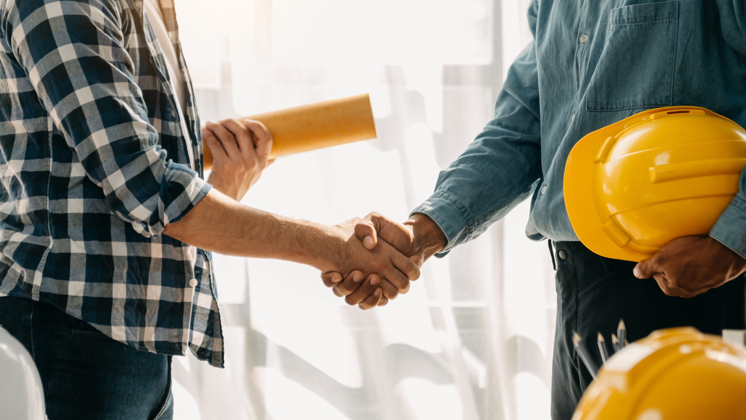 Construction team shake hands Two people shaking plans, one holds rolled up project plans and the other holds a safety helmet.