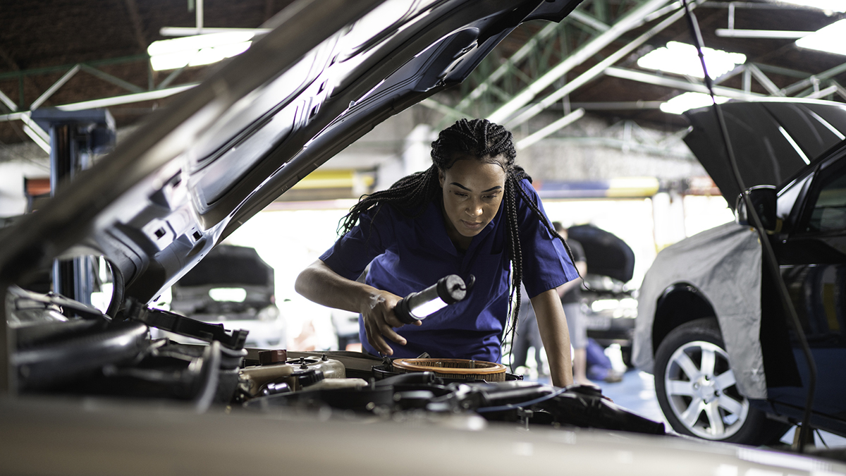 An automobile service worker inspects and engine compartment with a light.