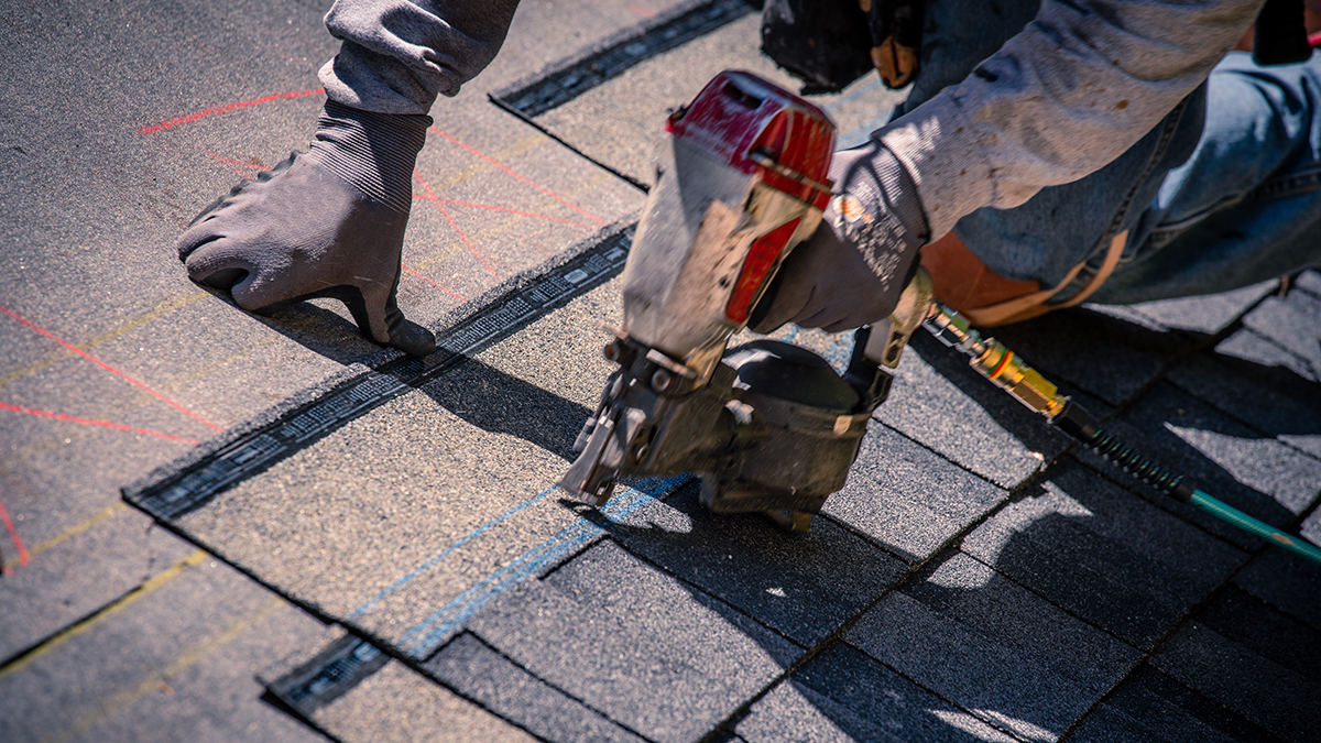 Someone's hands using a nail gun on a roof to install shingles