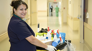 Adult cleaner with cleaning cart in a hospital hallway.