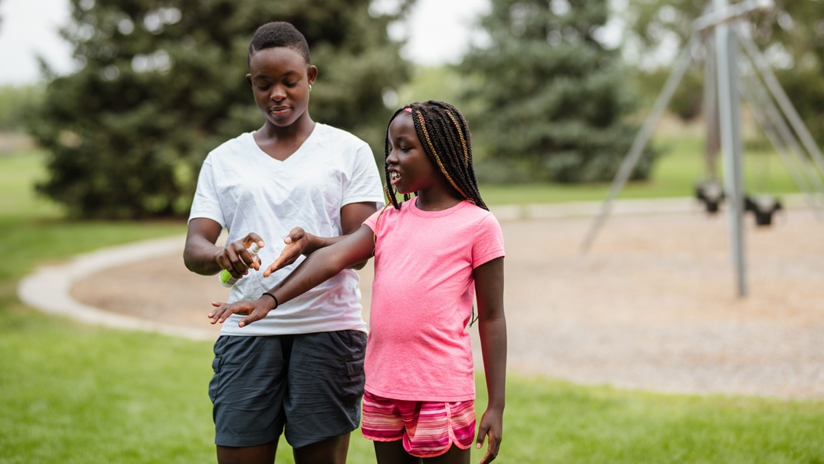 An adult man applies insect repellent to a young girl's arm in a park.