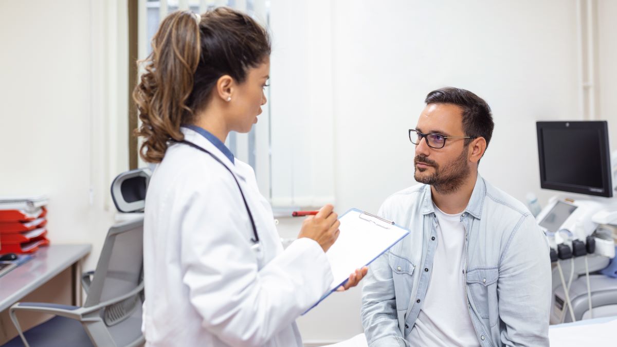 Female doctor stands as she talks to a sitting male patient in an exam room.