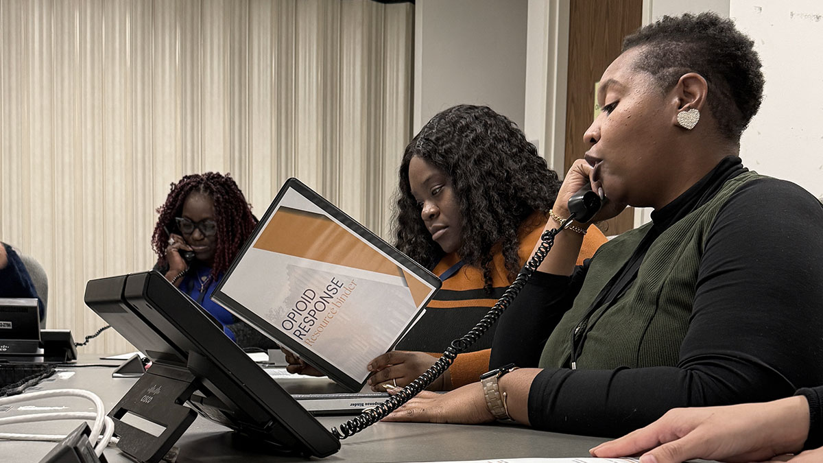Three women sitting at a conference table with phones and a handbook answering calls to the Overdose Prevention Hotline