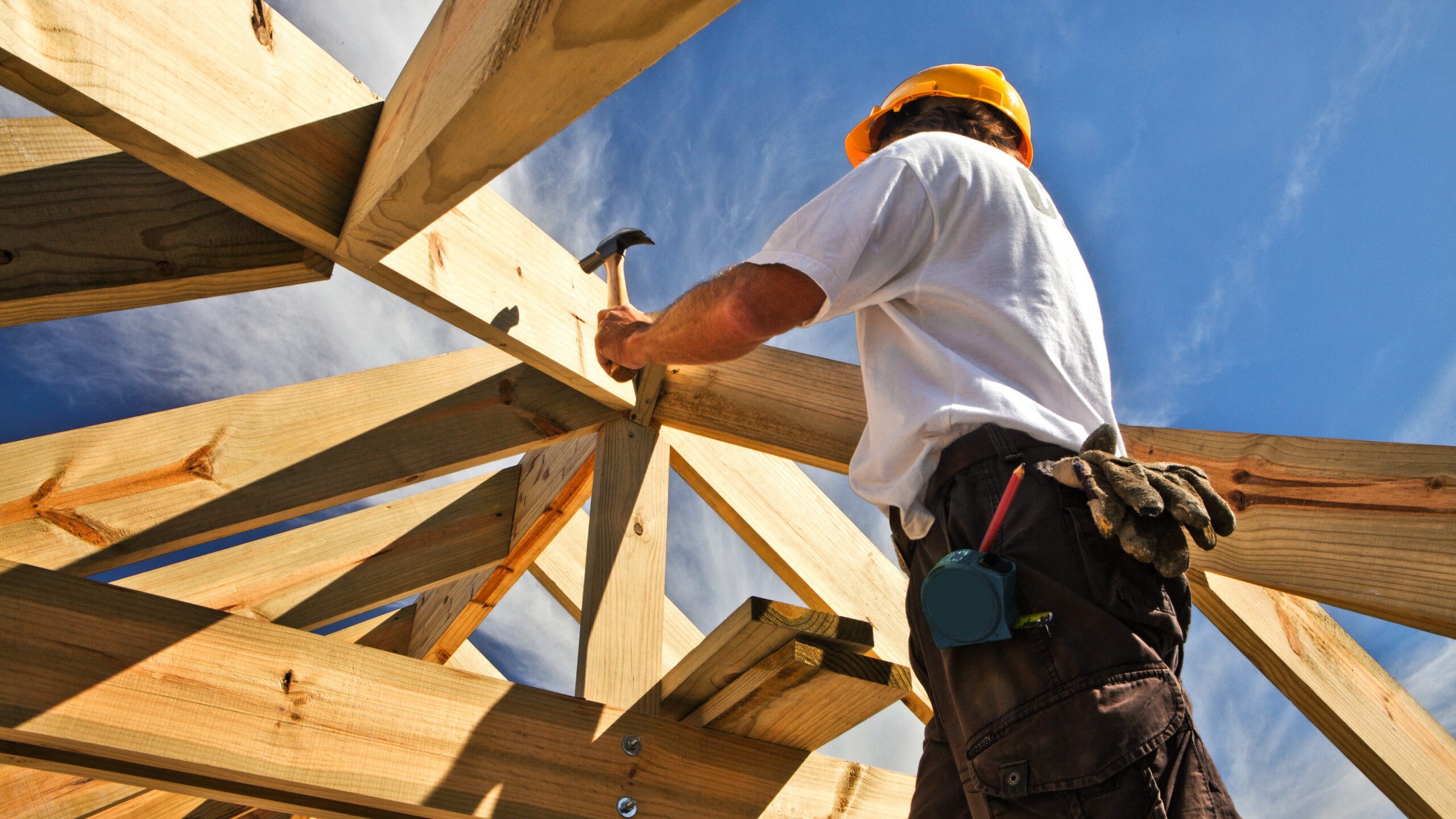 A roofer working on the wooden framing of a roof with blue sky in the background.