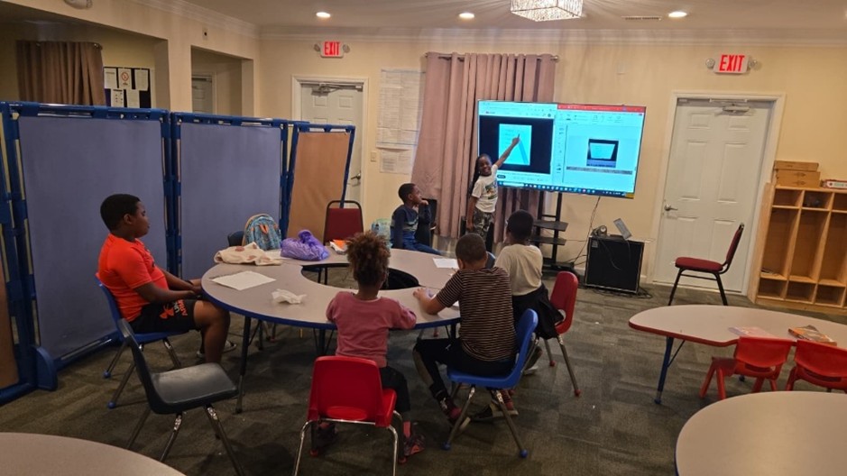 A group of children sitting around a table and one child standing and speaking in front, pointing to a screen.