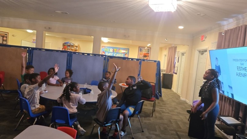 Group of children watching another child present, with some raising their hands
