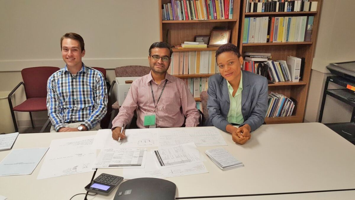 Three people sitting a large table strewn with papers in an office setting with bookshelves.