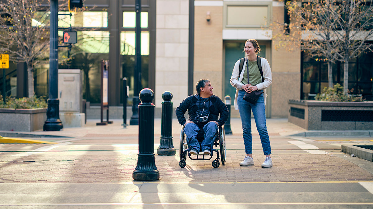 maninwheelchairyoungerwomanstandinginfrontofbuildinggetty.jpg Man in wheelchair and woman standing about to cross the road.