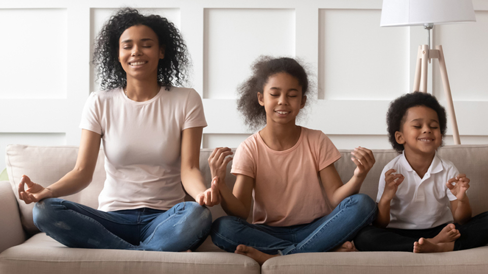 stay calm Young black mother sitting cross-legged on a couch with her two small children sitting similarly next to her in meditation.