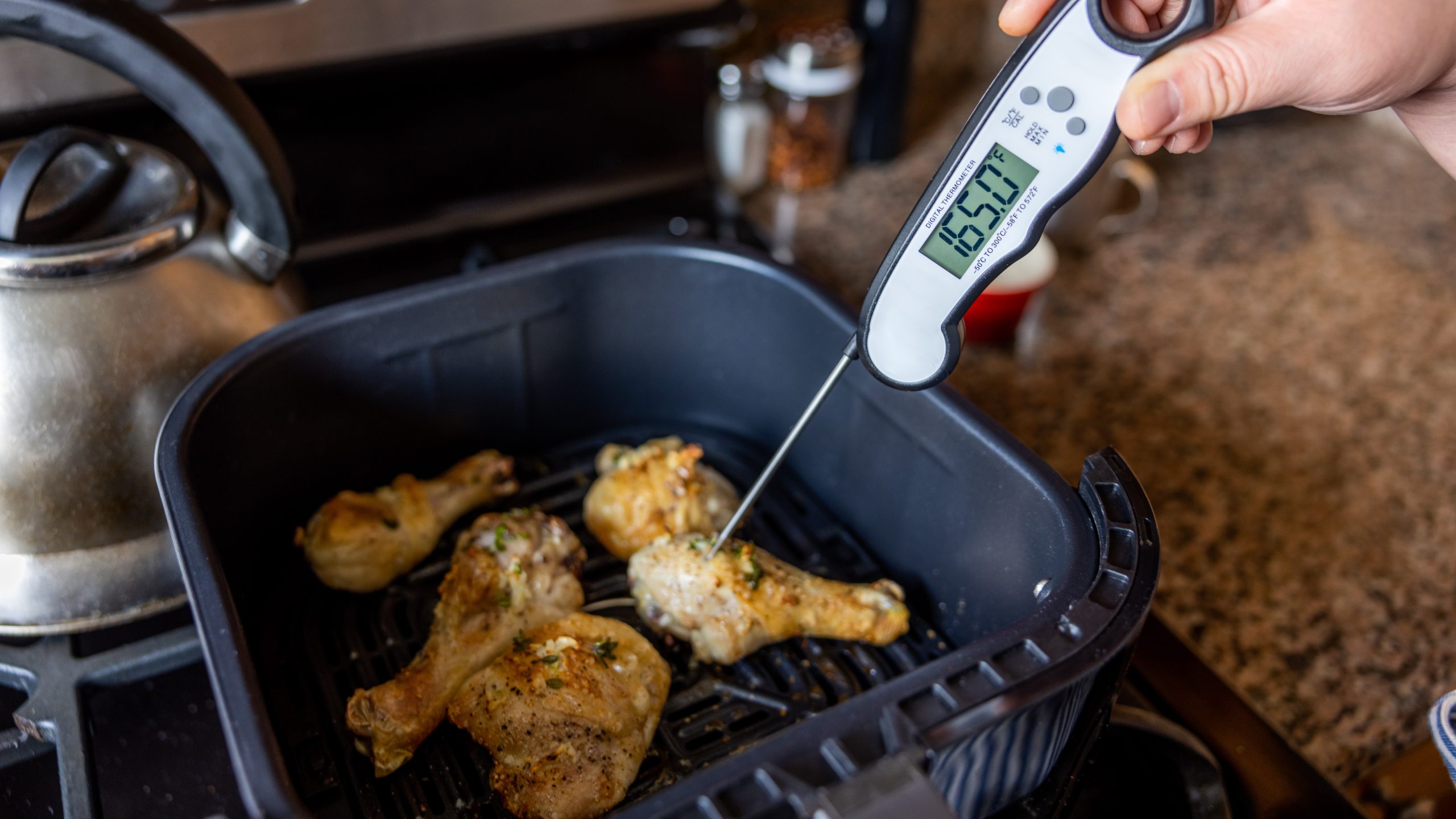 Person inserting food thermometer into chicken pieces in an air fryer to check temperature. Thermometer reads 165&deg;F.