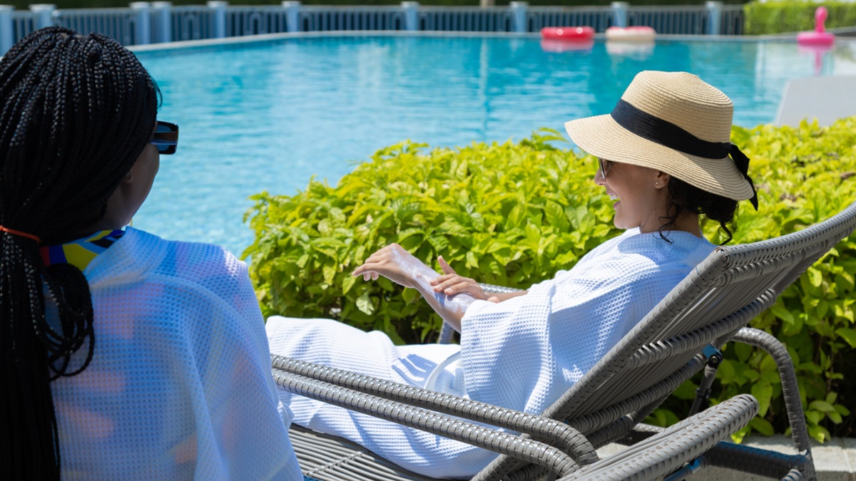Two women sitting near a pool. Both are wearing sunglasses. One is wearing a hat and putting sunscreen on her arm.