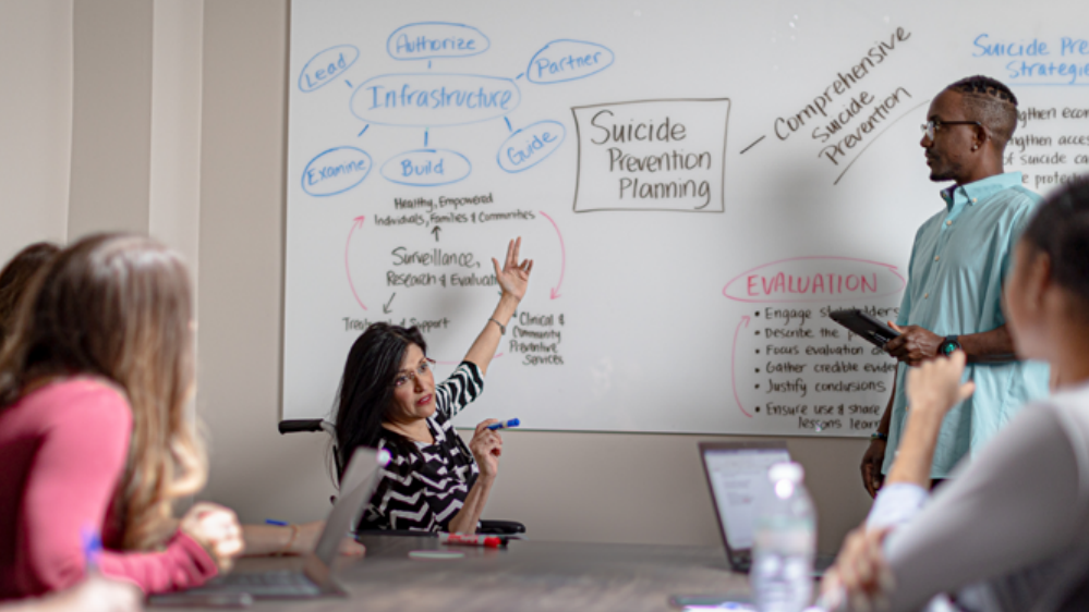 A group of people gathered around a table talking and writing on a whiteboard.
