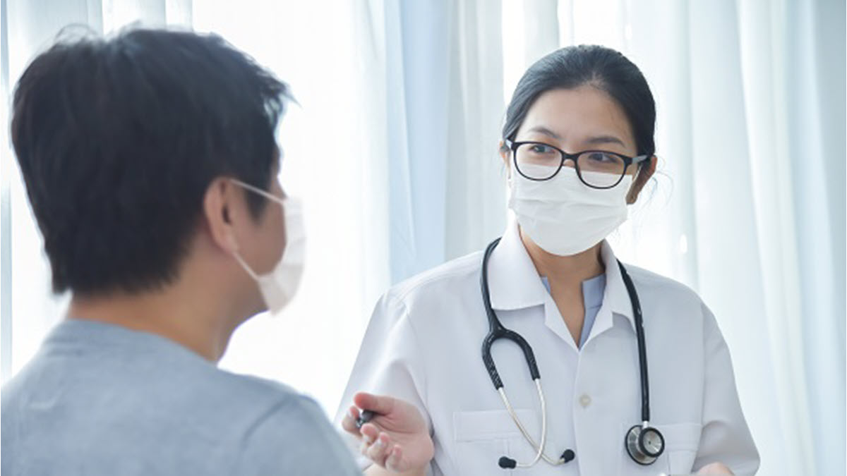Una m&eacute;dica hablando con un paciente; ambos est&aacute;n usando mascarillas.