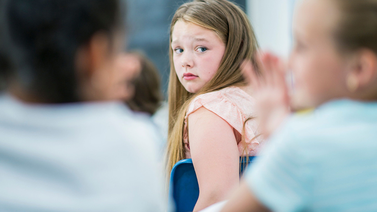 Two young girls making fun of another student their age in a classroom.