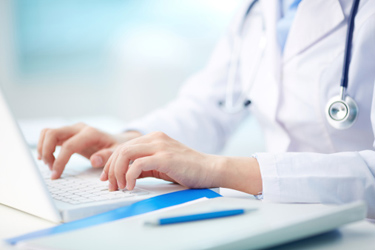 A close-up of a healthcare professional&rsquo;s hands typing on a laptop keyboard. The person is wearing a white coat with a stethoscope around their neck.