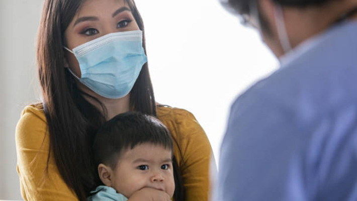 Masked mom holds her child in front of a doctor at an appointment.