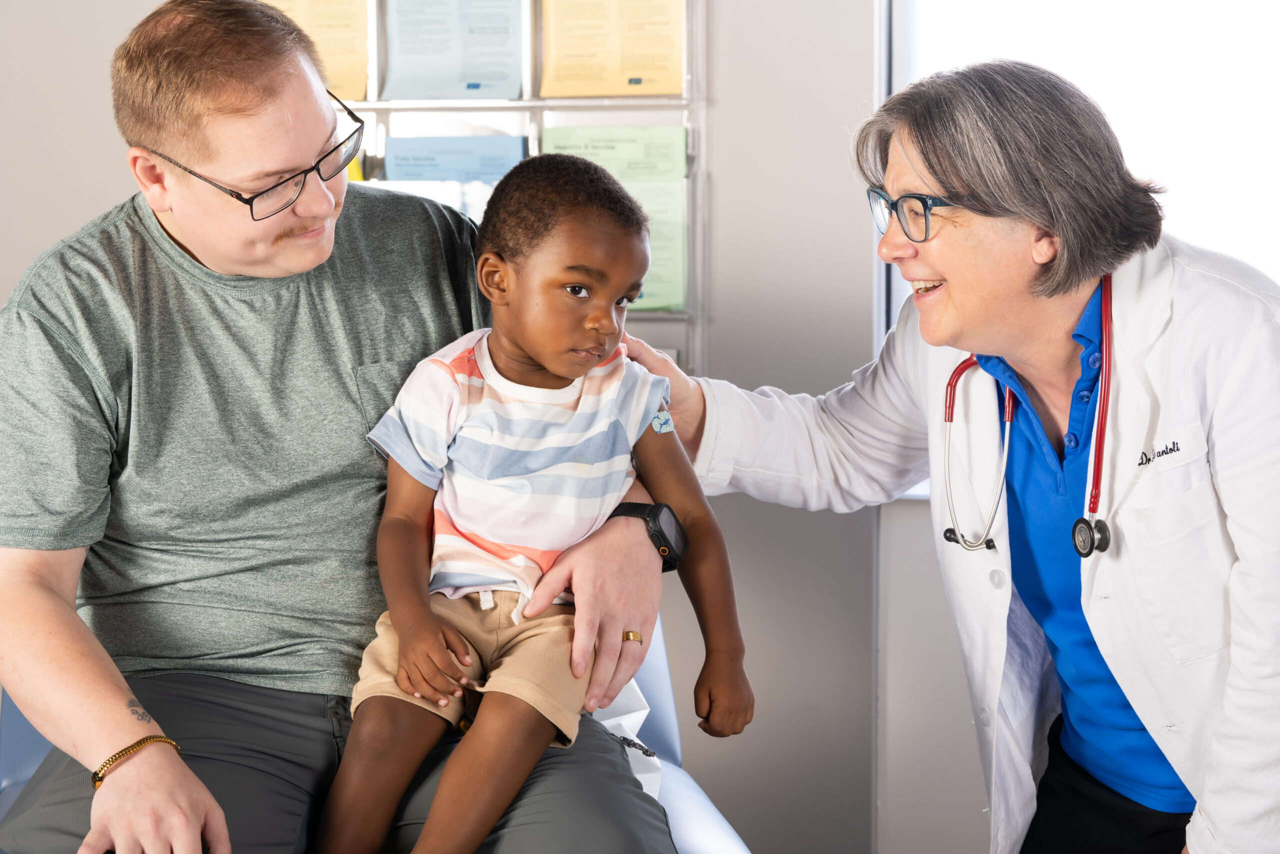 A child wearing a band-aid while being attended to by a caregiver and a pediatrician.