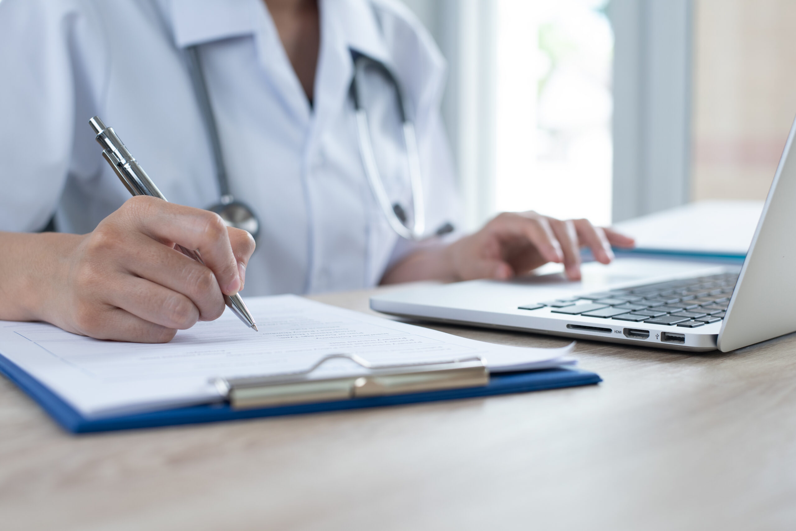 Doctor looking at computer and writing on paper attached to a clipboard