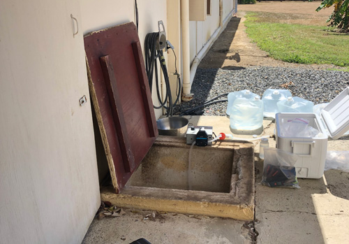 Una cisterna subterr&aacute;nea externa al lado de una edificaci&oacute;n. La tapa de madera est&aacute; abierta y se ve la abertura de concreto cuadrada. Hay una nevera port&aacute;til, bolsas de agua y un instrumento para pruebas del agua alrededor de la abertura de la cisterna.