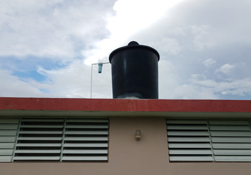 Edificaci&oacute;n con un recipiente de almacenamiento de agua grande (cisterna) en el techo. Hay un tubo de pl&aacute;stico entre la cisterna y el techo, con un filtro de agua conectado al tubo.