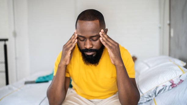 Man holding his hands to his head while sitting on the edge of a bed.